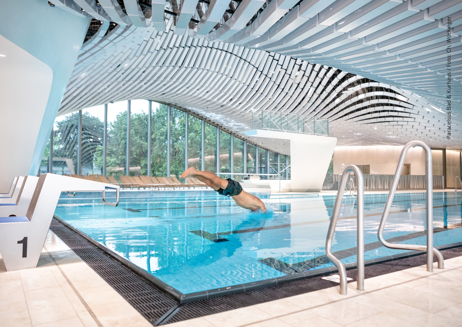 View through an indoor swimming pool. A man jumps head first into the pool. Text: "Paracelsus Bad und Kurhaus / Foto: Ch. Wöckinger"